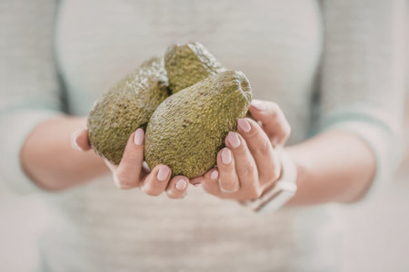 Woman holds three fresh green avocados in her hands, showcasing a soft pink manicure and a relaxed...