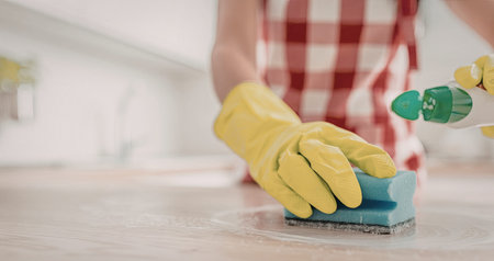 Person wearing green gloves and red checkered apron cleans a wooden surface with a sponge and spray...