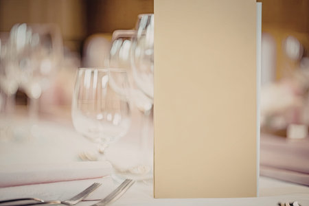 Fine dining table setting with blank yellow menu card and wine glasses on white tablecloth in...