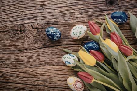 Decorated easter eggs and a bunch of vibrant red, yellow tulips arranged artfully on a rustic wooden...