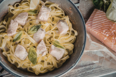 Freshly cooked salmon pasta with basil leaves and lemon in a metal pan on a wooden table ready to be...