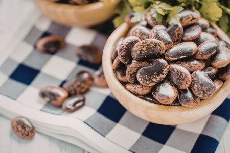 Speckled beans are piled high in a wooden bowl on a blue and white checkered tablecloth.