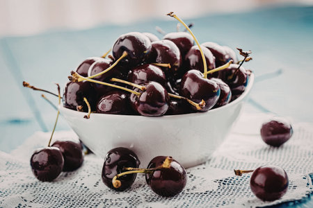 Fresh dark cherries overflowing from a white bowl placed on a white lace cloth with some cherries scattered around.の写真素材