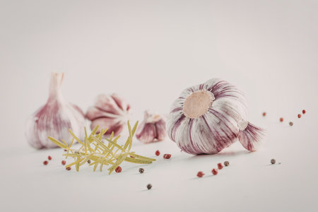 Fresh garlic bulbs and rosemary herb accompanied by scattered peppercorns on a white surface...