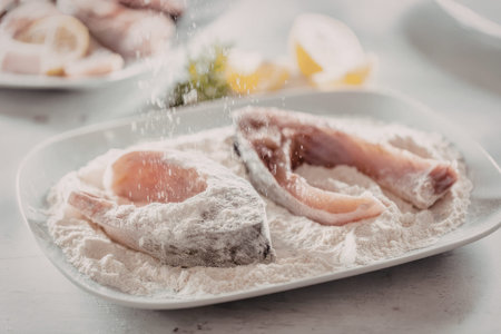 Fresh raw fish fillets are being coated with flour on a white plate preparation for a delicious...