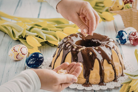 Person decorates easter cake with chocolate glaze and colorful sprinkles surrounded by tulips and...