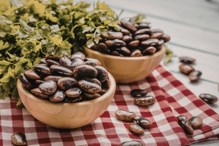 Speckled beans fill two wooden bowls on a red and white checkered tablecloth with fresh parsley and...