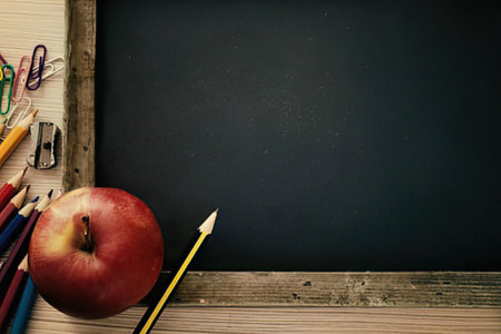A still life image of a red apple, a yellow pencil, and a bunch of colored pencils and pens on a wooden desk in front of a blank blackboard. The blackboard has a wooden frame and is mounted on a beige wall. The overall scene suggests a classroom or educational setting.