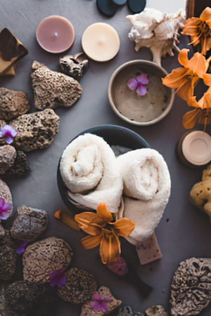 a peaceful spa arrangement featuring rolled towels in a bowl surrounded by rocks purple flowers and lit candles in various colors on a gray surface with orange leaves and a bowl of sand with a flower