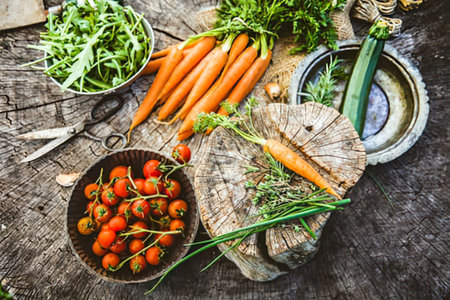 An overhead shot of a selection of fresh vegetables and greens arranged on a rustic wooden table. The vegetables include carrots, cherry tomatoes, and a zucchini, all accompanied by various herbs and greens. A wooden cutting board holds some of the produce, while metal bowls and a pair of scissors are also visible.