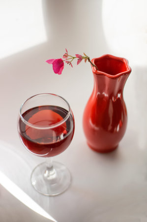 Still life with a pink flower in a bright red vase and a glass of wine on a blurred background