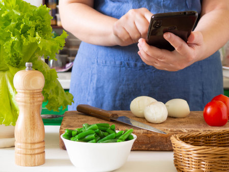 A faceless cook in an apron prepares a nicoise salad on the kitchen table and looks at the phone for a clue. Food blogger takes photos in the kitchen.の写真素材