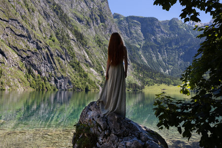 A young woman stands on a stone near a mountain lake of emerald colorの写真素材