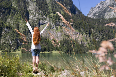 A slender red-haired long-haired woman stands on the shore of a clear lake in the Alpine mountainsの写真素材