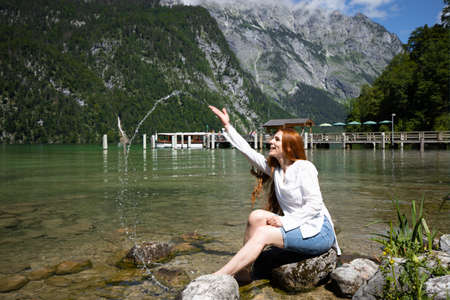 A slender red-haired beautiful woman sits on the shore of a clear mountain lake and plays with splashesの写真素材