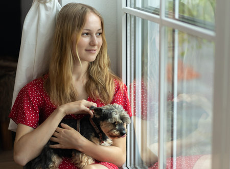 A romantic girl in a red dress sits on the floor near the window, strokes a dog and looks out the...