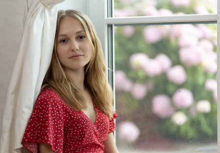 A young girl in a red dress sits on the floor in front of a bush with pink flowers and looks at the cameraの写真素材