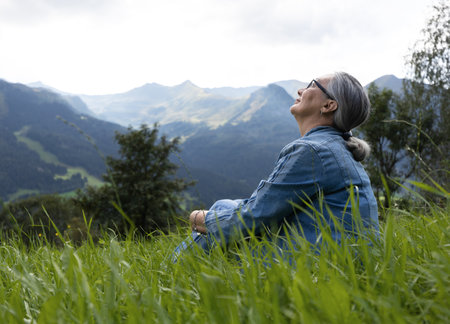 An elderly woman in a denim suit sits on a alpine meadow and looks at the skyの写真素材