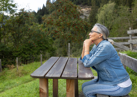 A gray-haired woman sits at a wooden table in the countryside at the edge of the forest with a kind look.の写真素材