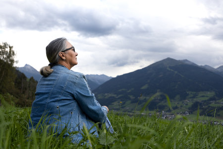 An elderly woman in a denim suit sits on an alpine meadow and looks towards the mountains.の写真素材