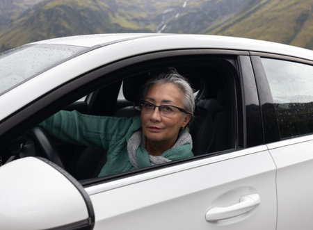 A gray-haired lady sits behind the wheel of a passenger car with an open window and looks at the cameraの写真素材