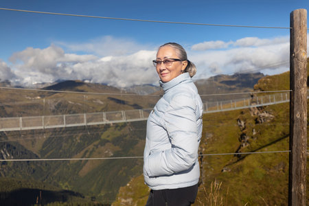 A woman in sportswear stands at the fence against the background of a suspension metal bridge in the Austrian Alpsの写真素材