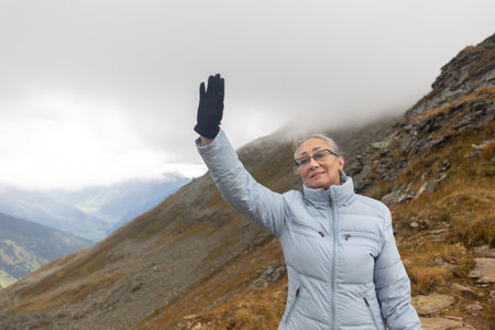 A gray-haired woman in sportswear stands at the top of a mountain and waves her hand in a friendly manner. Austria, Alpsの写真素材