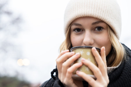 A young girl in a woolen hat drinks a hot drink from a mugの写真素材