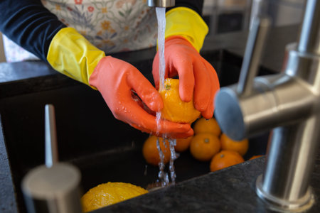 Womens hands in bright gloves wash orange fruit with tap water