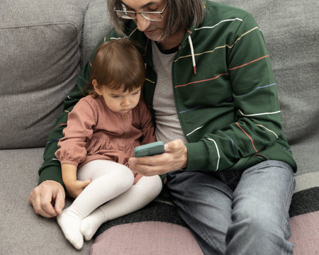 Grandfather and granddaughter sit on the home couch and look at the mobile phone