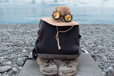 A backpack, a straw hat with glasses and an old sneaker lie by the seashoreの写真素材