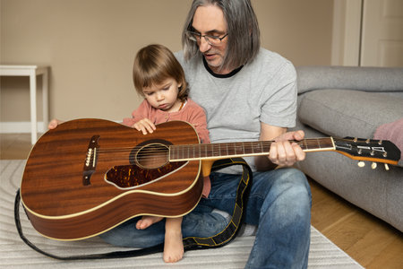 A grandfather introduces a baby granddaughter to a guitar while sitting on the floor.の写真素材