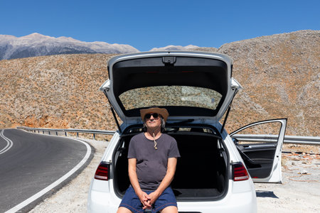 Male traveler sits at the open trunk of a car parked on the road uphill, Greece, Creteの写真素材