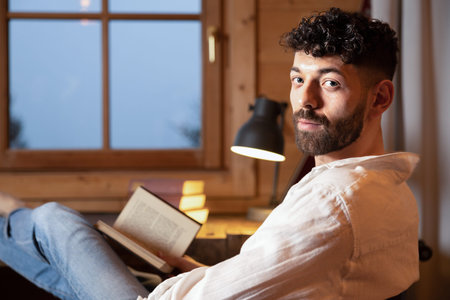 Young curly-haired man sitting at home with a book in his hands against the background of a window