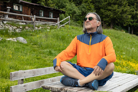 A long-haired gray-haired man sits on a wooden table in a yoga pose against the backdrop of old abandoned farm, Austriaの写真素材