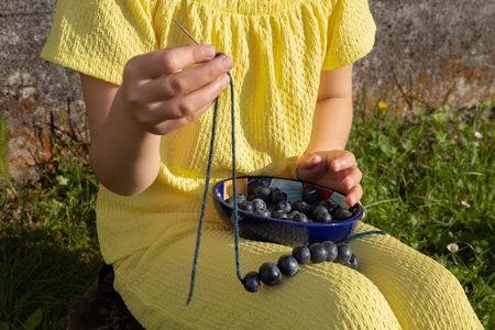 A girl in a bright yellow suit sits on the lawn and collects blueberry beadsの写真素材