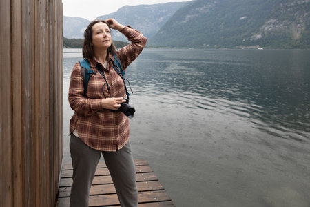 Young female photographer standing on the shore of lake with camera in her hands and looking into the distance, Austriaの写真素材