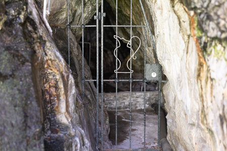 The entrance to the cave with source of mineral water closed with a metal lattice. On the grate there is a jug for waterの写真素材