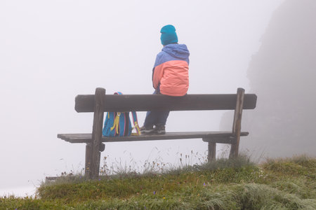 Little girl sitting with her back on a bench in the mountains on a foggy day, Austriaの写真素材