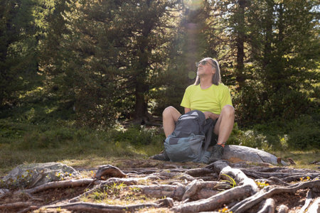 An adult man with a backpack sits in a sun-drenched clearing with tree rootsの写真素材