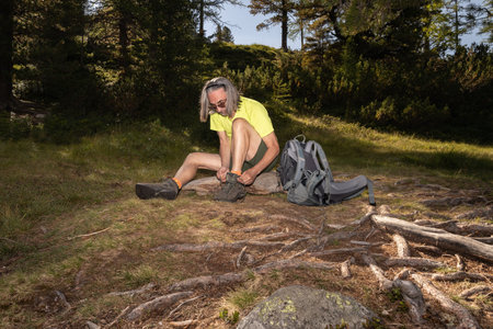 An adult gray-haired man with a backpack sits at the roots of a tree and ties a shoelace in the evening lightの写真素材