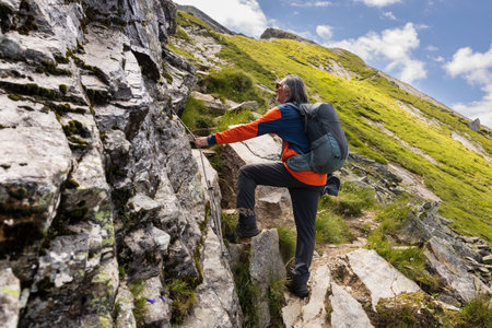 Gray-haired long-haired man with backpack climbing up the slope to the summit through Sala with railing, Austriaの写真素材