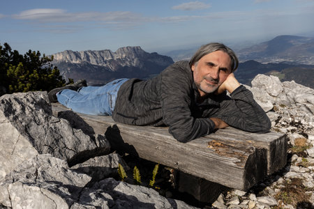 Gray-haired smiling man lying on a wooden bench on the background of a picturesque mountain landscape, Austriaの写真素材