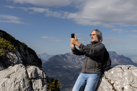 Gray-haired long-haired man with backpack taking selfie on steep slope on picturesque mountain background, Austriaの写真素材