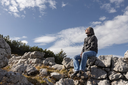 Adult gray-haired man sitting on a rocky fence against a cloudy sky and looking at the clouds, Austriaの写真素材