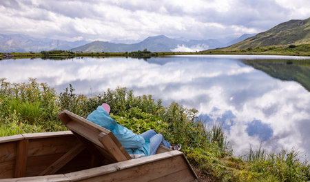A woman in the wind sits on the shore and admires a picturesque mountain lake with a reflection cloud, the conceptの写真素材