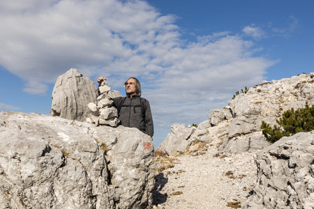Gray-haired long-haired man building a tower of stones against the background of a picturesque sky and a rocky mountainの写真素材