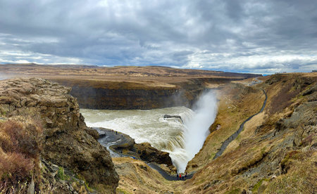 A picturesque Icelandic landscape with a powerful waterfall, a red slope and a dramatic skyの写真素材