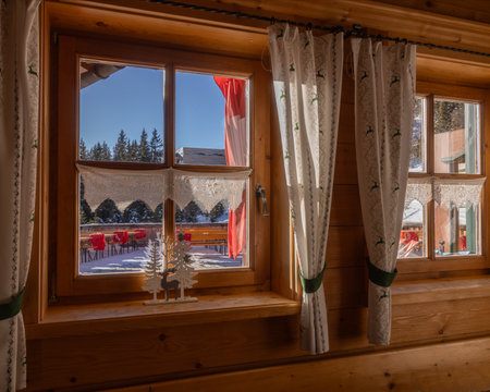 Snowy winter view through a cozy wooden cabin window in a serene alpine location with holiday decorationsの写真素材