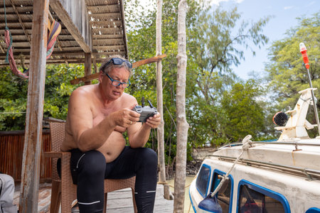 Adult man operates a drone while sitting by a boat in a tropical setting during daytimeの写真素材